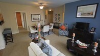 This single-floor residence features an open-concept layout that seamlessly connects the living and dining areas into one cohesive, functional space. The living area is anchored by a white sofa adorned with decorative pillows in navy and gray tones, accompanied by a dark ottoman and accent chair upholstered in a patterned fabric with an orange throw pillow. A bold slate-blue accent wall adds visual depth and contrast to the otherwise neutral, warm-toned interior. The dining area accommodates a dark wood table surrounded by white upholstered chairs, set atop a patterned area rug. A decorative centerpiece with tall glass vases and candlesticks adds an elegant touch to the dining setup. The ceiling fan with integrated lighting provides both airflow and illumination throughout the shared space. Soft, wall-to-wall carpeting runs throughout, contributing to a warm and comfortable ambiance. Framed artwork decorates the walls, adding a curated, polished feel. A hallway visible in the background leads to additional rooms, suggesting a thoughtful and efficient floor plan throughout the residence.