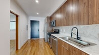 Located at 1525 N Hobart Blvd in the heart of Los Angeles, this modern 2-bedroom, 2-bathroom unit offers a thoughtfully designed single-level layout with clean contemporary finishes throughout. Light-toned hardwood floors run the length of the space, complementing crisp white walls and recessed lighting to create a bright, cohesive aesthetic.

The kitchen is a standout feature, showcasing rich walnut-toned cabinetry that extends floor to ceiling, paired with marble-style quartz countertops and a subway tile backsplash. Stainless steel appliances — including a gas range, microwave, dishwasher, and French door refrigerator — complete the functional yet stylish space. A striking dark front door adds a bold design accent at the entry.

The open living area leads to a glass-door balcony, allowing natural light to filter through and providing views of the surrounding neighborhood, including palm-tree-lined streets. Both bedrooms are finished with the same warm hardwood flooring and feature black-framed windows that frame outdoor views. The primary bedroom includes a mirrored sliding closet, accessed through a distinctive slatted wood barn-style door.

The two bathrooms are finished with neutral tile flooring and marble-look surrounds. One features a glass-enclosed walk-in shower, while the other offers a full tub-shower combination with sliding glass doors and grab bars.