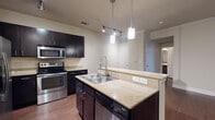 This single-story residence is designed with a practical, open layout that connects key living areas seamlessly. The kitchen serves as the centerpiece of the home, showcasing dark espresso cabinetry that extends to the ceiling, offering generous storage throughout. Granite countertops with warm, neutral tones run across both the perimeter surfaces and the central island, which features a double-basin stainless steel sink with a flexible gooseneck faucet and pendant lighting suspended above. A full suite of stainless steel appliances includes a French door refrigerator, electric range, built-in microwave, and dishwasher. Wood-look flooring runs through the kitchen and living areas, adding warmth and visual continuity. Track lighting and pendant fixtures ensure the space is well-lit throughout. The bedroom offers plush carpeting underfoot, a ceiling fan, and a window that brings in natural light. An attached bathroom features a soaking tub with tile surround, wood-look flooring, and grab bars for added convenience. The overall ambiance is clean and modern, with neutral tones and functional finishes throughout the home.