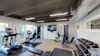 This fitness center offers a thoughtfully designed, single-floor exercise facility built to accommodate a wide range of workout needs. The space features a modern aesthetic defined by light wood-look flooring, crisp white walls, and a bold dark exposed-beam ceiling fitted with linear LED lighting that ensures bright, even illumination throughout.

The cardio zone is well-stocked with treadmills, elliptical trainers, a recumbent bike, and a rowing machine, all positioned along large windows that flood the area with natural light and provide pleasant outdoor views of surrounding greenery. Protective rubber floor mats are placed beneath each piece of equipment for safety and comfort.

The strength training area includes a multi-station cable machine by Hoist, an adjustable weight bench, and a dumbbell rack, providing versatile options for resistance training. A wall-mounted flat-screen television is centrally positioned for entertainment during workouts.

Glass partition walls on one side of the gym offer a visual connection to an adjacent lounge or common area, contributing to an open, airy feel. Exposed ductwork and HVAC systems are integrated into the ceiling design, adding an industrial touch. The overall layout is spacious and uncluttered, allowing for easy movement between equipment zones and making this fitness center a fully functional and inviting environment for daily exercise.