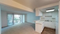 This single-floor unit offers a clean, functional layout designed for comfortable everyday living. The open-concept living and kitchen area serves as the heart of the space, where a peninsula-style kitchen counter seamlessly connects both zones. The kitchen is well-appointed with white cabinetry offering generous storage, light countertops, a gas range, built-in microwave, refrigerator, and a double sink — all finished in a cohesive white palette. Wood-look flooring grounds the kitchen area, while plush carpeting extends throughout the living and bedroom spaces, adding warmth underfoot. The living area benefits from a large sliding glass door fitted with vertical blinds, inviting natural light and providing views of lush greenery outside. A structural beam detail adds architectural character to the ceiling, which features a textured finish throughout. The bedroom is a spacious, light-filled retreat with neutral walls and carpeted flooring, complemented by an exterior door that introduces additional natural light. The overall ambiance is bright, airy, and move-in ready, with a neutral color scheme that provides a versatile backdrop for a variety of interior styles.