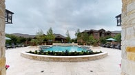 This expansive outdoor pool area is designed with a resort-style aesthetic, centered around a generously sized swimming pool with clear blue water and a smooth stone coping surround. The pool features integrated spa areas and entry steps, framed by raised limestone retaining walls adorned with agave plants, ornamental grasses, and flowering shrubs that provide year-round visual interest. A travertine tile deck extends throughout the space, offering ample room for sunbathing and socializing. Multiple seating arrangements are distributed across the deck, including cushioned lounge chairs, shaded conversation sets with large umbrellas, and additional dining-height tables positioned along pathways. Mature palm trees and tall ornamental trees contribute to the lush, tropical ambiance. A covered outdoor kitchen area with a built-in stainless steel grill is situated beneath a rustic wood pergola attached to a limestone-clad clubhouse building, which serves as an architectural focal point. The overall design balances natural materials—stone, wood, and greenery—with upscale amenities, creating a refined and inviting outdoor retreat.