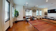 Located at 1951 Old Haywood Rd in Asheville, NC, this single-floor community space is thoughtfully designed to accommodate a variety of activities and social interactions. The open layout is unified by warm-toned flooring throughout, creating a cohesive and inviting atmosphere.

The main gathering area features a full-sized ping-pong table as a central focal point, complemented by a stylish lounge nook with upholstered bench seating, accent pillows, and bistro-style tables. Two sculptural woven globe pendant lights hang overhead, adding a decorative yet functional lighting element. Multiple windows dressed with sheer white curtains and horizontal blinds bring in generous natural light and views of surrounding greenery.

A dedicated entertainment wall showcases a large flat-screen TV mounted against a textured stone tile accent wall, flanked by a white cabinetry unit providing countertop and storage space. Adjacent to this area, a business center offers built-in computer workstations and a communal high-top table with bar stools, making it a practical workspace. Recessed lighting throughout ensures the entire space remains well-lit and functional at all hours.