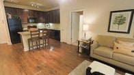 This single-floor space offers a thoughtfully designed open-concept layout that seamlessly connects the kitchen, dining, and living areas into one cohesive and functional environment. Rich, warm hardwood flooring flows throughout, adding a sense of continuity and elegance to the entire space.

The kitchen is outfitted with dark espresso cabinetry that extends to the ceiling, providing ample storage while creating a bold, sophisticated aesthetic. A classic tile backsplash complements the cabinetry, and the kitchen is equipped with full appliances including a black range and microwave. A raised breakfast bar with bar stool seating serves as a natural divider between the kitchen and the open living area, making it ideal for casual dining or entertaining.

The living area is warm and inviting, with soft ambient lighting provided by a classic table lamp. Crown molding along the ceiling adds an architectural detail that elevates the overall finish of the space. A doorway with clean white trim leads to additional areas of the home, hinting at further private accommodations beyond.

Overall, the space strikes a balance between practicality and style, with its dark cabinetry, hardwood floors, and crown molding details contributing to a comfortable and refined atmosphere.