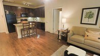 This single-floor space offers a thoughtfully designed open-concept layout that seamlessly connects the kitchen, dining, and living areas into one cohesive and functional environment. Rich, warm hardwood flooring flows throughout, adding a sense of continuity and elegance to the entire space.

The kitchen is outfitted with dark espresso cabinetry that extends to the ceiling, providing ample storage while creating a bold, sophisticated aesthetic. A classic tile backsplash complements the cabinetry, and the kitchen is equipped with full appliances including a black range and microwave. A raised breakfast bar with bar stool seating serves as a natural divider between the kitchen and the open living area, making it ideal for casual dining or entertaining.

The living area is warm and inviting, with soft ambient lighting provided by a classic table lamp. Crown molding along the ceiling adds an architectural detail that elevates the overall finish of the space. A doorway with clean white trim leads to additional areas of the home, hinting at further private accommodations beyond.

Overall, the space strikes a balance between practicality and style, with its dark cabinetry, hardwood floors, and crown molding details contributing to a comfortable and refined atmosphere.
