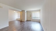 This two-floor residence offers a functional and clean layout spread across two levels, connected by a carpeted staircase with a white handrail. The main level greets you with a generously sized open living area finished in durable wood-look flooring that extends throughout the home. Large windows dressed with vertical blinds allow natural light to fill the space, complementing the neutral white walls and creating a bright, airy atmosphere. A ceiling fan overhead adds comfort to the living area, which flows openly into an adjacent room for flexible use. The galley-style kitchen is outfitted with warm wood cabinetry, dark countertops, a white appliance suite including a refrigerator and range, and a stacked washer and dryer unit — combining practicality with efficient use of space. The upper level hosts well-proportioned bedrooms, each featuring the same wood-look flooring, ample wall space, and windows that bring in natural light. One bedroom includes a sliding closet and direct access to an adjoining bathroom. The bathroom is finished with white cabinetry, a single vanity with a mirrored medicine cabinet, and tile flooring. An additional storage area beneath the staircase provides useful extra space. Overall, the home presents a clean, move-in ready environment with a straightforward, efficient design.