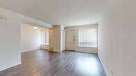 This two-floor residence offers a functional and clean layout spread across two levels, connected by a carpeted staircase with a white handrail. The main level greets you with a generously sized open living area finished in durable wood-look flooring that extends throughout the home. Large windows dressed with vertical blinds allow natural light to fill the space, complementing the neutral white walls and creating a bright, airy atmosphere. A ceiling fan overhead adds comfort to the living area, which flows openly into an adjacent room for flexible use. The galley-style kitchen is outfitted with warm wood cabinetry, dark countertops, a white appliance suite including a refrigerator and range, and a stacked washer and dryer unit — combining practicality with efficient use of space. The upper level hosts well-proportioned bedrooms, each featuring the same wood-look flooring, ample wall space, and windows that bring in natural light. One bedroom includes a sliding closet and direct access to an adjoining bathroom. The bathroom is finished with white cabinetry, a single vanity with a mirrored medicine cabinet, and tile flooring. An additional storage area beneath the staircase provides useful extra space. Overall, the home presents a clean, move-in ready environment with a straightforward, efficient design.