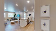 This single-floor residence is designed with a clean, contemporary aesthetic that prioritizes open living and functional layout. The heart of the home is an open-concept kitchen and living area connected by light wood-tone flooring and recessed lighting overhead. The kitchen is well-appointed with white shaker cabinetry, a large quartz-topped island offering bar seating for several guests, a black matte faucet, an undermount sink, and a full suite of stainless steel appliances including a refrigerator, range, and microwave. The island serves as a natural divider between the kitchen and the adjoining living space.

The living area is bright and welcoming, featuring multiple windows that allow natural light to fill the room and offer views of the surrounding neighborhood. The space comfortably accommodates a sofa arrangement and entertainment setup.

The bedrooms are carpeted and serene, each benefiting from natural light through sizable windows. One bedroom features ample space for a desk area, making it suitable for a home office setup as well.

The bathroom is finished with wood-look flooring and includes a walk-in glass-enclosed shower with bold black framing, lending a modern, upscale touch to the space. Overall, the unit presents a cohesive, move-in-ready environment with neutral tones throughout.