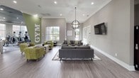 Located at 1420 W McDermott Dr in Allen, Texas, this single-story amenity center offers a thoughtfully designed collection of spaces for fitness, work, and relaxation. The facility is unified by a contemporary aesthetic featuring neutral gray tones, dark tray ceilings, wood-look flooring, and abundant natural light from large windows throughout.

The fitness area is expansive and well-appointed, divided into distinct zones. A cardio and strength room is lined with floor-to-ceiling windows and equipped with treadmills, ellipticals, rowing machines, and free weights. An adjacent studio space features spin bikes, freestanding punching bags, and balance equipment, with large wall mirrors and ceiling fans enhancing the workout environment.

A welcoming lounge area anchors the social hub of the facility, complete with plush gray and green seating, a globe pendant chandelier, wall-mounted televisions, and a bold green accent wall with "GYM" lettering. Nearby, a stylish seating area with black egg-style chairs and mounted screens provides a more intimate gathering spot.

The business center offers individual computer workstations with pendant lighting and dark wood column accents. A private conference room features a solid wood table, green upholstered chairs, geometric pendant lights, and expansive windows with outdoor views — ideal for meetings or collaborative work.