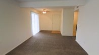 This single-floor space features an open living area finished with wall-to-wall dark carpeting that extends throughout the main room. A large floor-to-ceiling window dressed with vertical blinds allows natural light to filter into the space, creating a bright and airy atmosphere. A ceiling-mounted light fixture with a gold-toned base provides additional overhead illumination. The front entry door is centrally positioned along the main wall, with a small hard-surface landing that transitions into the carpeted living area. Neutral white walls keep the space feeling clean and versatile. An open passageway on the right side of the living area leads to additional rooms, suggesting a functional layout that connects the main living space to the rest of the unit. The architectural design is straightforward and practical, with a structural beam detail visible along the ceiling that subtly defines the entry zone from the broader living area. Overall, the space offers a blank canvas well-suited for customization, with its neutral finishes and simple layout providing flexibility for a variety of furniture arrangements and design preferences.