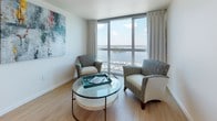 This single-floor waterfront residence is defined by its expansive floor-to-ceiling windows that frame breathtaking panoramic views of a marina and river beyond. Natural light floods the living areas, where plush carpeting and neutral tones create a calm, sophisticated ambiance throughout.

The living space is generously proportioned, offering multiple seating arrangements that take full advantage of the stunning water and marina views. Whether used as a lounge or a den, the room's large-format windows ensure the scenery is always the focal point.

The kitchen is efficiently designed with warm wood cabinetry, granite countertops, a glass cooktop, stainless steel dishwasher, and an over-the-range microwave — providing all the essentials for comfortable living. Light hardwood-style flooring adds warmth to the space.

The bedroom is a serene retreat, featuring soft carpeting, generous natural light from corner windows, and a tranquil atmosphere well-suited for rest. Ample space accommodates a full furniture arrangement with ease.

The bathrooms are finished to a high standard, with large-format tile flooring, wood vanity cabinetry, a white countertop, and a glass-enclosed walk-in shower with tasteful tile surround — delivering a clean, spa-like aesthetic throughout.