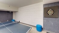 This energetic game room is a well-appointed recreational space spread across a single floor, offering a variety of entertainment options in a compact yet efficiently organized layout. The room is defined by its bold design choices, most notably its striking red accent walls that create a lively, dynamic atmosphere complemented by crisp white walls and a painted white brick feature wall that adds texture and visual depth.

Light wood-look flooring runs throughout, providing a clean, modern foundation that ties the space together. The game room is equipped with a full-size ping-pong table, a foosball table, a classic shuffleboard table, and a dartboard — offering diverse recreational options for guests or residents. Blue cylindrical stools provide casual seating near the ping-pong table, while a pair of bold red lounge chairs offer a comfortable spot to relax between games.

Colorful wall art, including an abstract multicolored painting and a graphic vertical bar mural in blue, orange, and grey, adds artistic flair to the space. A workstation counter with pendant lighting is tucked into one corner alongside a mini bar area with an illuminated refrigerator. The room is well-lit with a combination of flush circular ceiling fixtures and linear fluorescent lighting, and includes a wall-mounted mini-split HVAC unit for climate control.
