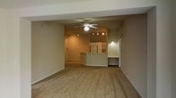 This single-floor residence is thoughtfully designed around an open-concept floor plan that seamlessly connects the living, dining, and kitchen areas. The spacious living area is finished with wall-to-wall carpeting in a neutral beige tone, creating a warm and cohesive foundation throughout the space. A ceiling fan with an integrated light fixture is centrally positioned, providing both comfort and ambient illumination.

The kitchen is a standout feature, highlighted by a curved peninsula island that provides additional counter space and serves as a natural divider between the kitchen and living areas. White cabinetry lines the kitchen walls, and track lighting is installed overhead, ensuring the workspace is well-lit. Appliances, including a refrigerator, are visible and integrated into the cabinetry layout.

Adjacent to the kitchen, a recessed built-in desk alcove offers a dedicated workspace or study nook, complete with overhead lighting and shelving — a practical and efficient use of space. Track lighting is also present in the main living area, lending the room a flexible and modern lighting scheme.

The overall ambiance of the space is functional and understated, with neutral tones, soft carpeting, and purposeful built-in features that contribute to a comfortable, move-in-ready environment.