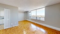 Located on an upper floor at 2405 Finch Avenue W in Toronto, this one-bedroom, one-bathroom unit offers a well-organized layout across a single level. Warm parquet flooring runs throughout the living spaces, adding a classic character to the interior. The open living room is generously proportioned and benefits from expansive floor-to-ceiling windows that frame sweeping panoramic views of the Toronto skyline and surrounding cityscape, filling the room with abundant natural light. The kitchen is tucked adjacent to the living area and features white cabinetry, a tile floor, and white appliances, offering a clean and functional workspace. The bedroom is a comfortable, well-lit space with its own window overlooking the city views, continuing the parquet flooring and neutral grey tones found throughout. A closet provides practical storage within the bedroom. The bathroom is finished with full-height white tile surrounding a tub-shower combination, a toilet, and a small window for ventilation. Neutral grey walls and white trim are consistent throughout the unit, contributing to a clean, cohesive aesthetic. The textured ceilings and baseboard heating are characteristic features of the building's design.