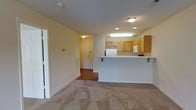 This single-floor residential space offers a practical and comfortable layout designed for everyday living. The open-concept design seamlessly connects the living area to the kitchen, creating a spacious and airy feel. The living area features light neutral carpeting that adds warmth underfoot, while the entryway transitions to hardwood-style flooring, providing a clear distinction between spaces.

The kitchen is well-appointed with warm medium-toned wood cabinetry offering ample storage, white appliances including a refrigerator and range, and recessed as well as under-cabinet lighting that ensures a bright and functional workspace. A peninsula-style breakfast bar extends from the kitchen, serving as both a casual dining space and a natural divider between the kitchen and living areas.

Soft, neutral yellow-toned walls run throughout the unit, contributing to a bright and inviting atmosphere. The textured ceiling adds a subtle architectural detail, while flush-mount and recessed lighting fixtures ensure consistent illumination across all areas. The entryway, visible from the main living space, features a solid entry door and provides a clean transition into the home. Overall, the space presents a cohesive, functional design with a warm and livable ambiance.