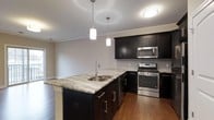 This single-floor space showcases a well-appointed kitchen as its centerpiece. The kitchen is designed with rich, dark-toned cabinetry that extends to the upper and lower sections, providing generous storage throughout. A large kitchen island with granite countertops serves as a functional and visual focal point, complete with an undermount stainless steel sink and a sleek, modern faucet. Two pendant lights hang above the island, casting warm, directed lighting over the workspace.

The countertops feature a striking granite surface with natural veining, complementing the dark cabinetry. Stainless steel appliances, including a gas range, over-the-range microwave, and dishwasher, add a contemporary touch. A subway tile backsplash lines the walls behind the range, contributing texture and visual interest.

Crown molding trims the ceiling, adding an elegant architectural detail throughout the space. The warm hardwood flooring grounds the kitchen and flows cohesively into the adjacent areas. The neutral wall tones and ambient lighting create a clean, inviting atmosphere throughout.