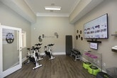 This single-floor fitness studio is thoughtfully designed to support a variety of workout styles in a clean, organized environment. The space features wood-look flooring throughout and neutral gray-toned walls, creating a calm yet motivating atmosphere. Crown molding and tray ceiling details add a polished architectural touch, while recessed and flush-mounted lighting ensures the space is well-lit throughout.

The cycling area accommodates multiple stationary bikes arranged with ample spacing, allowing for group or individual sessions. A "Breathe" wall decal and decorative gear-themed accents contribute to the room's fitness-focused aesthetic. An adjacent workout zone is equipped with a rack of red dumbbells in varying weights, step platforms, resistance tools, and a wire shelving unit for organized equipment storage.

A large multi-pane window allows natural light to filter in and offers views of an outdoor area, enhancing the open feel of the space. A wall-mounted flat-screen TV with interactive fitness content provides on-demand workout guidance. Additional amenities include wall-mounted shelving for accessories, a coat hook rail, and easy access to an adjoining room, likely a restroom or changing area, rounding out the facility's practical offerings.