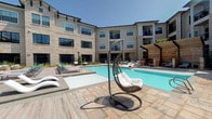 This outdoor pool area offers a well-appointed retreat designed for relaxation and recreation. The centerpiece is a large, clearly maintained swimming pool with calm, blue-green water, complemented by an adjacent spa. The expansive pool deck is finished with stone-toned pavers and wood-look decking, providing ample space for lounging. Seating options are varied and plentiful, including teal chaise lounges, freestanding swing chairs, and additional seating tucked beneath a wood-framed pergola structure, lending a cabana-style atmosphere to one section of the deck.

The pool is nestled within a courtyard framed on multiple sides by a multi-story residential building featuring stucco and stone facade detailing, large windows, and private balconies. Mature trees and landscaped greenery soften the hardscape and provide natural shade throughout the area. Access to the pool is provided through a dedicated entry point marked by rustic wood-framed doors and posted pool rules signage.

The overall ambiance is bright and open, benefiting from abundant natural sunlight and clear sky views. The design balances modern architectural elements with warm, natural materials, creating a cohesive and inviting outdoor living environment suitable for both active use and leisurely relaxation.
