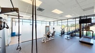 Located at 1500 SW Aileron Cir in Bentonville, Arkansas, this professional fitness facility offers a thoughtfully designed exercise space spanning a single floor. The layout is divided into distinct functional zones, each tailored to a specific type of training.

The cardio area is anchored by a row of treadmills and elliptical machines, each equipped with individual monitors for an engaging workout experience. Floor-to-ceiling glass walls line the perimeter, flooding the space with natural light and offering open views of the surrounding outdoor greenery, creating an energizing atmosphere.

A dedicated strength training zone features a power rack, adjustable bench, and a full dumbbell rack, providing everything needed for resistance-based workouts. The durable rubber flooring in this area ensures safety and stability during heavy lifts.

A separate functional training area transitions to light hardwood-style flooring and includes stability balls, medicine balls, battle ropes, and a stationary bike — ideal for dynamic, multi-modal workouts. Glass partition walls define this zone while maintaining visual openness across the facility.

The drop ceiling with recessed panel lighting ensures consistent, bright illumination throughout. The overall design balances a clean, modern aesthetic with practical, performance-driven functionality, making this a versatile and well-appointed fitness destination.