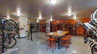 This single-floor bike room is thoughtfully designed to accommodate cycling enthusiasts with both storage and maintenance functionality. The space features an extensive system of wall-mounted bicycle racks lining the perimeter, maximizing storage capacity while keeping bikes organized and easily accessible. A freestanding double-sided floor rack in the center of the room provides additional storage for multiple bikes simultaneously.

At the heart of the room sits a sturdy butcher block worktable supported by industrial-style pipe legs, surrounded by matching orange metal stools. This central station serves as a dedicated bike repair and maintenance area, offering ample surface space for tools and equipment.

The room's design is characterized by bold orange accent walls that complement the orange seating, creating a cohesive and energetic aesthetic. The polished concrete flooring is both durable and easy to maintain, well-suited for a high-traffic utility space. Flush-mount ceiling light fixtures provide consistent, even illumination throughout. The overall layout is practical and efficiently organized, making it an ideal amenity for cyclists seeking secure storage and a convenient workspace.