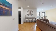 This well-appointed single-floor space offers a functional and modern layout, combining a kitchen and living area within an open-concept design. The living room is bright and welcoming, featuring warm hardwood-style flooring, recessed lighting, and crisp white walls that create a clean, contemporary ambiance. A wall-mounted flat-screen TV anchors one side of the room, while a large sectional sofa and a stylish bar-height console table with stools provide comfortable seating and social space. A distinctive oversized gear-style clock serves as a striking decorative focal point on the wall. Large windows allow natural light to filter in, enhancing the sense of openness. The kitchen is efficiently designed with a sleek, galley-style layout featuring floor-to-ceiling white cabinetry, gray countertops, a stainless steel double-basin sink, and modern hardware throughout. A microwave and refrigerator complete the kitchen's practical amenities. The bathroom is thoughtfully designed with accessibility in mind, equipped with multiple stainless steel grab bars, a paper towel dispenser, and light tile flooring. The space is rounded out by exit and emergency signage, indicating its suitability for shared or communal use. Overall, the space exudes a clean, modern aesthetic with practical, well-considered finishes.