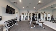 Located at 7200 Chadwood Lane in Dublin, Ohio, this single-floor fitness center is designed to support a comprehensive workout experience. The space is laid out across an open, elongated floor plan finished with durable, gray carpet-style flooring suited for exercise use throughout.

The strength training area is prominently featured and includes a multi-station cable machine, a rack with weight plates and barbells, and multiple adjustable benches. A tiered dumbbell rack holds a full range of free weights and is positioned beneath a wall-mounted flat-screen television, offering entertainment during workouts. Full-length mirrors line one wall, visually expanding the space and aiding in form correction.

The cardio section is situated toward one end of the room and houses treadmills, an elliptical, and a stationary bike. Motivational wall decals, including phrases like "Sweat," "Fat Burning," and "Beast Mode On," add an energizing atmosphere to the environment. Framed motivational posters reinforce the theme.

Natural light enters through large windows and glass-panel doors, offering views of the surrounding outdoor area. Recessed lighting supplements illumination throughout. A water cooler, sanitizer dispenser, and ventilation system round out the practical amenities, ensuring comfort during use.