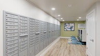 This community mailroom is a thoughtfully designed, functional space that serves as a central hub for residents. The room is dominated by a large, floor-to-ceiling bank of numbered, individual gray mailboxes lining the full length of one wall, providing organized and secure mail access for a sizable number of residents. The light wood-look flooring runs throughout, complementing the clean white walls and recessed lighting that illuminates the space evenly. At one end, a soft sage-green accent wall adds a touch of warmth and visual interest, adorned with a framed abstract artwork. A small lounge area nearby features casual blue seating options arranged atop a patterned area rug, creating a brief respite for residents. The opposite end of the space includes a compact kitchenette with white cabinetry, a countertop coffee station, a sink, and a wall-mounted flat-screen television, enhancing convenience and comfort. Package storage shelving and additional utility features are also present. Multiple doorways connect the space to adjacent areas, and the overall layout is open and easy to navigate, reflecting a clean, modern aesthetic designed with community functionality in mind.