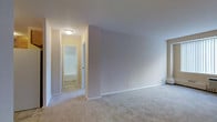 This single-floor unit is laid out in an L-shaped configuration, providing a clear separation between the living area and the bathroom and storage spaces. The main room is generously sized, finished with wall-to-wall carpeting in a neutral beige tone that adds warmth underfoot. The walls are painted in soft, light hues, contributing to a bright and airy atmosphere. Flush-mount ceiling lighting illuminates the space evenly, and the room includes at least one window that allows natural light to filter in. A hallway connects the main living area to a full bathroom, which includes a bathtub and toilet, finished with tiled flooring. Adjacent to the hallway is a closet, offering dedicated storage space. The overall layout is compact yet functional, with a straightforward flow between areas. The clean, neutral palette throughout provides a versatile backdrop suitable for a variety of uses. The unit appears to be vacant and move-in ready, with all surfaces in presentable condition.