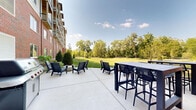 Located at 77 S Adams St in Oswego, Illinois, this outdoor common area offers a well-appointed exterior space designed for community gathering and relaxation. The building itself is a modern multi-story structure, featuring a combination of classic red brick and contemporary gray siding, with private balconies visible along its facade.

The centerpiece of the outdoor area is a generous concrete patio furnished with Adirondack-style chairs in a deep charcoal finish, high-top tables with bar seating, and a round concrete fire pit table — creating multiple zones for socializing and lounging. A gas grill station is also available, making the space well-suited for outdoor entertaining.

Surrounding the patio, meticulously maintained lawns stretch broadly alongside the building, bordered by young trees, evergreen shrubs, and thoughtfully placed landscaping. The grounds open up to a wide, gently sloping greenspace that provides a park-like atmosphere and ample room for outdoor activities.

The mature trees lining the perimeter offer natural shade and a sense of seclusion, while the open sky above and lush surroundings create a bright, welcoming ambiance. The overall setting balances modern architectural design with natural greenery, offering residents a comfortable and attractive outdoor environment in a suburban Oswego setting.
