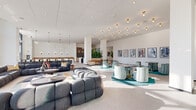 Located at 811 Peachtree St NE in Atlanta's Midtown neighborhood, this generously sized single-floor amenity space offers a sophisticated, contemporary environment designed for both relaxation and productivity. The layout spans a substantial footprint and is organized into several distinct functional zones that flow seamlessly into one another.

The main lounge area is a showstopper, featuring floor-to-ceiling windows that frame panoramic views of the Atlanta skyline and flood the space with abundant natural light. Oversized, sculptural seating arrangements — including tufted modular sofas and rounded accent chairs — are complemented by brass pendant and ceiling-mounted light fixtures that add warmth to the crisp white palette. A curated gallery wall of framed artwork adds a creative, cultural touch throughout.

A café-style bar area is outfitted with a white quartz island, woven counter stools, patterned tile flooring, and a sleek wet bar backed by dark ceramic tile. Adjacent seating with high-top tables and lounge furniture creates an inviting social hub. A recreational zone features a foosball table alongside casual seating with direct city views through expansive glazing.

Private glass-walled office spaces offer transparency and natural light while maintaining acoustic separation. A wide hallway corridor with glass partitions connects the various zones, maintaining an open and airy feel throughout the entire floor.