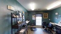 This single-floor salon space makes a strong first impression with its rich teal-painted walls, which lend a bold, professional character to the interior. The space is designed to accommodate styling and hair care services efficiently, featuring at least one dedicated styling station complete with a wall-mounted mirror, a white countertop for tools and products, and a professional salon chair positioned on a protective floor mat. A shampoo bowl station is visible toward the back of the room, equipped with a reclining chair, indicating full-service hair care capabilities.

Natural light filters into the space through a window dressed with a dark valance, complementing the overhead fluorescent lighting that ensures a well-lit working environment. The hardwood-style flooring adds warmth and is easy to maintain. A decorative metal shelving unit near the entrance displays salon products, a clock, and welcoming décor, reinforcing the space's organized yet personable atmosphere. Framed artwork on the walls adds a finishing touch. Overall, the space is thoughtfully arranged to balance functionality with a welcoming, boutique salon ambiance.