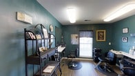 This single-floor salon space makes a strong first impression with its rich teal-painted walls, which lend a bold, professional character to the interior. The space is designed to accommodate styling and hair care services efficiently, featuring at least one dedicated styling station complete with a wall-mounted mirror, a white countertop for tools and products, and a professional salon chair positioned on a protective floor mat. A shampoo bowl station is visible toward the back of the room, equipped with a reclining chair, indicating full-service hair care capabilities.

Natural light filters into the space through a window dressed with a dark valance, complementing the overhead fluorescent lighting that ensures a well-lit working environment. The hardwood-style flooring adds warmth and is easy to maintain. A decorative metal shelving unit near the entrance displays salon products, a clock, and welcoming décor, reinforcing the space's organized yet personable atmosphere. Framed artwork on the walls adds a finishing touch. Overall, the space is thoughtfully arranged to balance functionality with a welcoming, boutique salon ambiance.