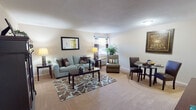 This single-floor studio in Kenner, Louisiana, offers a thoughtfully organized open-concept layout that maximizes every square foot. The main living area is carpeted and generously sized, accommodating a full seating arrangement, a dining area, and additional accent furniture without feeling cramped. Neutral-toned walls and plush carpet create a warm, inviting atmosphere throughout the space, while a window along one wall allows natural light to filter in and provides views of the outdoors.

A dedicated dining nook sits adjacent to the living area, offering space for a small table and chairs. The kitchen is positioned along one side of the unit and features warm wood cabinetry, a double-basin sink, standard appliances including a full-size refrigerator, and tile flooring — providing practical functionality in a compact footprint.

An entry corridor connects the main living space to the bathroom, which includes a tub-shower combination and vanity with mirror. The layout flows logically from entry to living and kitchen areas, making efficient use of the available space. Overall, the studio presents a clean, comfortable, and functional living environment suited for single occupancy.