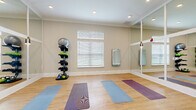 Located at 2150 NW 21st St in Ocala, Florida, this single-floor yoga and fitness studio offers a clean, purpose-built environment for exercise and wellness. The studio features warm hardwood-style flooring throughout, providing a practical and inviting surface ideal for yoga, stretching, and bodyweight training. Recessed ceiling lighting ensures even illumination across the entire practice area, while blinds-covered windows allow for natural light and privacy control.

A defining architectural feature is the large floor-to-ceiling mirrored wall framed in crisp white trim with glass panel dividers, which visually expands the space and allows practitioners to monitor their form during workouts. The mirrors open into an adjacent fitness area, creating a seamless connection between the two zones. Elegant French doors with frosted glass panels provide a refined transition between rooms.

Built-in bench seating and open cubbies near the entry offer convenient storage for personal belongings. A multi-tiered equipment rack along one wall holds stability balls, resistance bands, and kettlebells, keeping fitness tools organized and accessible. A smaller wall-mounted mirror is also present for localized viewing. The overall aesthetic is light, airy, and minimal, with neutral warm-beige tones contributing to a calm, focused atmosphere well-suited for both individual practice and group sessions.