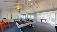 This single-story clubhouse in Gainesville, Florida offers a well-equipped recreational space designed for social gatherings and leisure activities. The interior is defined by striking vaulted ceilings with exposed white-painted beams, lending the space an open, airy feel. Woven pendant lights in warm yellow tones hang throughout, complementing the sage green accent walls and dark, textured carpeting that grounds the room.

The game area serves as the centerpiece of the clubhouse, housing a pool table, shuffleboard table, air hockey table, and foosball table—all featuring a consistent light wood and dark metal aesthetic. A ceiling fan circulates air overhead, adding to the comfort of the space.

Adjacent to the game area is a lounge zone with navy blue sofas arranged near a wall-mounted flat-screen television on a floating media console. This area provides a relaxed setting for casual viewing or socializing. Abundant windows along multiple walls admit generous natural light and offer views of the landscaped grounds. A water cooler and whiteboard are also present, adding functional utility. French doors provide direct access to the outdoor surroundings, further connecting the interior to the community outside.