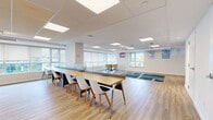 Located at 254 Phillip St in Waterloo, Ontario, this single-floor multipurpose space offers a generous, open-concept layout ideal for collaboration, study, or community gatherings. Light hardwood-style flooring runs throughout, creating a warm and cohesive aesthetic that complements the crisp white walls and drop-panel ceiling fitted with recessed LED lighting panels.

The space is thoughtfully organized into distinct functional zones. A lounge area features round tables paired with ergonomic yellow rolling chairs, positioned alongside roller-shade windows that invite soft, natural light and views of the surrounding neighbourhood. A dedicated study zone includes a long, partitioned workbench with individual white chairs, providing focused workstations for multiple users. An open lounge zone is anchored by a patterned area rug and a wall-mounted flat-screen TV atop a mid-century style media console.

Toward the rear of the space, a service counter with bar-style seating offers an additional casual work or social area, complemented by access to utility rooms and storage through multiple white interior doors. The overall ambiance is clean, modern, and welcoming — designed to support a variety of uses within a bright, well-appointed environment.