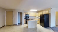 This single-floor residence offers a practical and well-organized layout designed for comfortable everyday living. The kitchen serves as a central highlight of the space, featuring a freestanding center island with a granite-style countertop that provides additional workspace and casual dining potential. Light wood cabinetry lines the walls, offering generous storage throughout, while black appliances — including a gas range, built-in microwave, refrigerator, and sink — add a sleek, cohesive look. The countertops carry a subtle stone-pattern finish, and the kitchen is illuminated by a ceiling-mounted fluorescent light fixture that ensures bright, even lighting. Light-toned tile flooring extends through the kitchen and into the adjacent areas, creating a seamless and easy-to-maintain surface. A closet with a built-in rod is conveniently accessible just off the kitchen, adding valuable storage to the layout. The neutral color palette throughout — warm beige walls paired with white trim — lends the space a clean, welcoming ambiance. The open sightlines between areas contribute to a sense of spaciousness, making the floor plan feel connected and airy throughout.