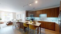 This well-designed single-floor residence combines modern aesthetics with functional living. The open-concept kitchen is a standout feature, showcasing rich wood-grain cabinetry that extends floor to ceiling, white quartz countertops, a subway tile backsplash with under-cabinet lighting, and stainless steel appliances. A central kitchen island with seating serves as both a dining and gathering space, flowing seamlessly into a bright living room with hardwood-style flooring and a large window that fills the space with natural light. The living area offers a comfortable, casual atmosphere with ample room for lounging and entertaining. Two well-appointed bedrooms each feature large windows overlooking the surrounding exterior, plush carpeting, and generous natural light. The primary bedroom connects to an en-suite bathroom featuring a glass-enclosed walk-in shower, tile flooring, and wood-grain vanity cabinetry with white countertops. A second full bathroom mirrors the same contemporary design language, complete with matching wood cabinetry and a large vanity mirror. Track lighting throughout the kitchen and common areas provides versatile illumination, while neutral wall tones create a clean, cohesive ambiance across the entire residence.