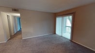 This single-floor space features a generously sized living area finished with wall-to-wall carpeting in a neutral tone, creating a comfortable and understated foundation. The room is framed by light beige walls and accented with clean white baseboards, contributing to a simple, functional aesthetic. A textured ceiling adds subtle visual interest overhead. The most notable feature of the living area is the full-length sliding glass door dressed with vertical blinds, which floods the space with natural light and offers a view of the surrounding greenery outdoors. The door provides direct access to an exterior area, enhancing the sense of openness. The layout is open and unobstructed, offering flexibility for a variety of furniture arrangements. Overall, the space presents a clean, neutral palette that serves as a versatile canvas for personalization.