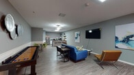 This single-floor communal space is thoughtfully designed to serve multiple functions, combining lounge, entertainment, work, and kitchen areas within one open, flowing layout. The interior is finished with light wood-look plank flooring and cool gray walls accented by a bold blue feature wall, creating a contemporary and inviting atmosphere throughout.

The lounge area anchors the space with a tufted blue sofa, modern accent chairs in tan leather and green upholstery, and a wall-mounted flat-screen television flanked by abstract artwork. A skylight introduces natural light, complementing the ceiling fans and recessed fixtures overhead. Adjacent to the lounge, a games area includes a shuffleboard table and a pool table, providing recreational options for residents or guests.

A dedicated workstation zone features multiple computer setups along the blue accent wall, illuminated by warm sconce lighting—suitable for focused work or study sessions. Nearby, a dining nook with a round table and colorful chairs offers a casual gathering spot.

The communal kitchen is well-appointed with stainless steel appliances, including a full-size refrigerator and range, white cabinetry with gray countertops, a subway tile backsplash, floating shelves, and a central island with bar stool seating—making it fully functional for group use.