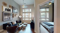 This single-floor residence makes a striking impression with its soaring ceilings and generous window arrangements that flood each room with natural light. The living area serves as the heart of the home, anchored by dramatic floor-to-ceiling window grids that frame outdoor views of the surrounding neighborhood. A tufted navy sofa, rustic wood coffee table, and curated gallery wall featuring framed artwork and woven decorative accents lend the space a sophisticated yet eclectic personality. Hardwood flooring flows throughout the main living areas, adding warmth and continuity.

The kitchen is well-equipped with rich espresso-toned cabinetry extending to the upper reaches of the tall walls, a distinctive wine rack detail, stainless steel appliances including a refrigerator, range, and microwave, and light quartz countertops complemented by a classic tile backsplash.

The bedroom is a tranquil retreat, featuring large multi-pane windows that invite ample daylight, soft carpeting, and a statement wall adorned with an arrangement of vintage bicycle wheels, adding a bold artistic touch. The bathroom continues the cohesive design with dark wood cabinetry, a wide framed mirror, and a tiled soaking tub and shower combination.