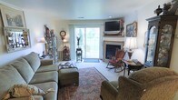 This single-floor residence offers a comfortable and thoughtfully arranged living space with a distinctly classic character. The living room serves as the heart of the home, centered around a traditional fireplace with a mantel that provides a natural focal point for the room. Above the fireplace, a mounted flat-screen television integrates modern convenience into the traditional setting. A sliding glass door fills the space with natural light and offers a view of the outdoors, creating a bright and airy atmosphere. The living room is furnished with plush, upholstered seating including a rocking chair, complemented by an ornate area rug that anchors the space and adds warmth underfoot. Decorative accents throughout — including a curio cabinet displaying collectibles, framed artwork, table lamps, and a mantel clock — contribute to the room's cozy, well-curated ambiance. The neutral-toned walls and light carpeting provide a clean backdrop that allows the furnishings and décor to stand out. Overall, the space conveys a welcoming and lived-in charm, ideal for relaxed everyday living and entertaining.