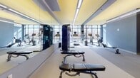 Located at 480 Rantoul Street in Beverly, Massachusetts, this fitness center offers a thoughtfully designed, single-floor workout facility suited for a wide range of exercise routines. The main gym area is bright and open, featuring floor-to-ceiling windows that flood the space with natural light and frame views of the surrounding landscape. The interior design is highlighted by bold yellow ceiling accents contrasted against steel-blue walls, creating an energizing atmosphere throughout. Full-length mirrors run along one wall, enhancing the sense of spaciousness and allowing users to monitor their form during workouts. The facility is equipped with a comprehensive array of cardio machines, including treadmills, stationary bikes, and rowing machines, as well as multi-function cable machines and adjustable weight benches. A dedicated free weights area features a tiered dumbbell rack paired with an adjustable bench, positioned beneath recessed and linear pendant lighting. A separate studio or flex room, finished with dark grey walls and durable flooring, provides space for yoga, stretching, or functional training, with equipment such as stability balls, foam rollers, and step platforms on hand. The overall layout is practical and well-organized, offering a complete fitness environment within a modern, polished setting.