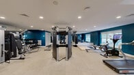 This fitness center offers a thoughtfully designed, single-floor exercise space that provides a comprehensive workout environment. The room is defined by striking deep teal accent walls contrasted against crisp white ceilings, creating a bold and energizing atmosphere. Recessed lighting throughout ensures the space is well-lit, while multiple large windows along the walls allow natural light to filter in and provide views of the surrounding greenery.

The cardio zone features several treadmills, elliptical machines, a stationary bike, and a rowing machine, many equipped with individual display screens. A dedicated functional training area includes a heavy bag, battle ropes, a RaX multi-functional rig, foam puzzle mats, and a medicine ball, catering to high-intensity and cross-training workouts.

The strength training section is well-appointed with a cable machine system, METAC functional trainers, a full dumbbell rack, and weight benches, all arranged to maximize floor space and usability. Full-length mirrors along one wall enhance the sense of openness and allow users to monitor their form. A water hydration station is conveniently located near the entrance. The light-toned flooring throughout ties the space together, delivering a clean, professional, and motivating fitness environment.