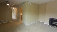 This single-floor residential space offers a functional and straightforward layout designed for comfortable everyday living. The main living area is finished with wall-to-wall carpet in a neutral beige tone, providing a soft and cohesive foundation throughout the space. The walls are painted in a warm, understated beige that contributes to a calm and inviting atmosphere.

Ceiling-mounted HVAC vents are visible, indicating a central air and heating system for climate control. The space flows naturally from the main living area into a hallway that connects to additional rooms, offering a practical and efficient floor plan. A glimpse into the kitchen reveals wood-toned cabinetry and what appears to be a countertop, suggesting a modest yet functional cooking and preparation area.

The hallway is well-lit and provides access to further areas of the unit, with a door visible at the far end, hinting at additional private spaces beyond. White baseboards run throughout, offering a clean contrast against the carpeted floors and painted walls.

Overall, this space presents a clean, move-in-ready environment with a neutral palette that allows for versatile interior styling. The layout is straightforward and practical, making efficient use of the available floor space across a single level.