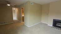 This single-floor residential space offers a functional and straightforward layout designed for comfortable everyday living. The main living area is finished with wall-to-wall carpet in a neutral beige tone, providing a soft and cohesive foundation throughout the space. The walls are painted in a warm, understated beige that contributes to a calm and inviting atmosphere.

Ceiling-mounted HVAC vents are visible, indicating a central air and heating system for climate control. The space flows naturally from the main living area into a hallway that connects to additional rooms, offering a practical and efficient floor plan. A glimpse into the kitchen reveals wood-toned cabinetry and what appears to be a countertop, suggesting a modest yet functional cooking and preparation area.

The hallway is well-lit and provides access to further areas of the unit, with a door visible at the far end, hinting at additional private spaces beyond. White baseboards run throughout, offering a clean contrast against the carpeted floors and painted walls.

Overall, this space presents a clean, move-in-ready environment with a neutral palette that allows for versatile interior styling. The layout is straightforward and practical, making efficient use of the available floor space across a single level.
