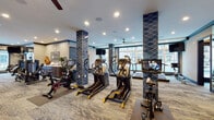 Located at 6480 Alexander Clay in Columbus, OH, this fitness center is a thoughtfully designed, single-floor facility that combines high-end equipment with an upscale aesthetic. The space is anchored by striking navy blue diamond-patterned tile columns that add a bold architectural statement throughout the main workout area. Recessed lighting and ceiling fans maintain a bright, comfortable environment, while large windows along the perimeter flood the space with natural light and offer views of the surrounding community.

The cardio zone is generously equipped with a full row of True-brand treadmills, multiple stair climbers, elliptical machines, and stationary cycling bikes, each featuring individual digital display screens. A dedicated strength training area houses a True multi-functional cable machine alongside isolated resistance machines, including a leg press and additional weight equipment, all set against textured patterned wall treatments with full-length mirrors to support proper form.

The flooring throughout combines durable carpet tile in neutral tones, providing comfort and traction underfoot. A separate enclosed studio space, visible through glass-paneled walls, offers additional room for focused training or stretching. The overall ambiance is polished and modern, reflecting a commitment to both functionality and design quality that elevates the everyday fitness experience.