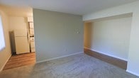 This single-floor residence offers a straightforward and functional layout designed for comfortable everyday living. The main living area is finished with plush carpeting in a neutral tone, providing a soft and inviting atmosphere. A smooth transition leads to adjacent areas finished with warm hardwood-style flooring, adding visual contrast and a sense of continuity throughout the space. The walls are painted in a soft, muted greige tone that lends a calm and neutral backdrop, well-suited to a variety of interior styles and furnishings. White painted baseboards frame the floor cleanly and contribute to the space's tidy, well-maintained appearance. An open passageway connects the living area to a secondary space, enhancing the flow and accessibility of the floor plan. The kitchen is visible from the main living area and features white appliances and cabinetry, maintaining the consistent, clean aesthetic found throughout. Recessed architectural details, including a built-in nook area with wood-panel accents, add a subtle design element to the space. The ceiling is smooth and white, contributing to a bright and open feel. Overall, the space presents a clean, neutral, and move-in-ready environment with a cohesive design throughout its single floor.