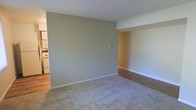 This single-floor residence offers a straightforward and functional layout designed for comfortable everyday living. The main living area is finished with plush carpeting in a neutral tone, providing a soft and inviting atmosphere. A smooth transition leads to adjacent areas finished with warm hardwood-style flooring, adding visual contrast and a sense of continuity throughout the space. The walls are painted in a soft, muted greige tone that lends a calm and neutral backdrop, well-suited to a variety of interior styles and furnishings. White painted baseboards frame the floor cleanly and contribute to the space's tidy, well-maintained appearance. An open passageway connects the living area to a secondary space, enhancing the flow and accessibility of the floor plan. The kitchen is visible from the main living area and features white appliances and cabinetry, maintaining the consistent, clean aesthetic found throughout. Recessed architectural details, including a built-in nook area with wood-panel accents, add a subtle design element to the space. The ceiling is smooth and white, contributing to a bright and open feel. Overall, the space presents a clean, neutral, and move-in-ready environment with a cohesive design throughout its single floor.
