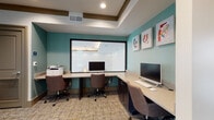 This business center is thoughtfully designed to accommodate a range of professional needs within a single, efficiently organized floor. The space is divided into two primary areas: an open workstation zone and a private meeting room accessible through glass-paneled double doors.

The workstation area features built-in desks along the walls, equipped with desktop computers and an HP multifunction printer, making it well-suited for tasks such as printing, scanning, and general computer use. The walls are painted in a calming teal tone, accented by colorful framed artwork that adds a vibrant touch to the workspace. A large interior window connects this area visually to the adjacent meeting room, enhancing the sense of openness.

The private meeting room offers a more focused environment, centered around a spacious wooden conference table surrounded by upholstered rolling chairs. Natural light filters in through tall windows dressed with elegant gray drapes, creating a calm and professional atmosphere. The interior window looking into the workstation area adds a layer of transparency between the two zones.

Throughout the space, patterned carpet flooring, warm taupe crown molding, and recessed lighting contribute to a cohesive and polished aesthetic. The overall design balances functionality with a welcoming ambiance, making it an effective environment for both collaborative meetings and individual work tasks.
